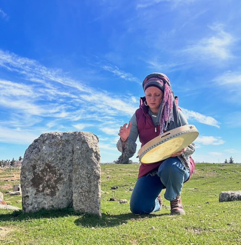 A woman kneeling with an Anatolian tambourine next to a broken foundation stone in the archaeological field at Harran, February 2024